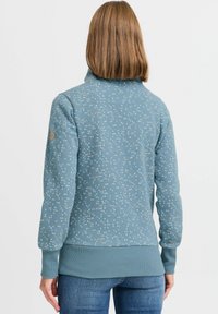 Sudadera de forro polar en azul con un patrón de pequeños puntos en rosa y blanco. Cuello alto, puños y dobladillo acanalados. Textura suave y corte casual.