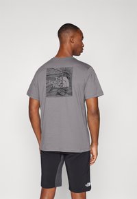 Pilkas trumpas rankovės marškinėliai su geometriniu juodu piešiniu ir "The North Face" logotipu nugaroje, derinami su juodais šortais.