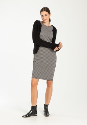 Schwarze Strickjacke über einem schwarz-weißen gemusterten Kleid. Das Kleid hat eine taillierte Form; das Model trägt schwarze Ankle Boots mit glänzender Oberfläche.
