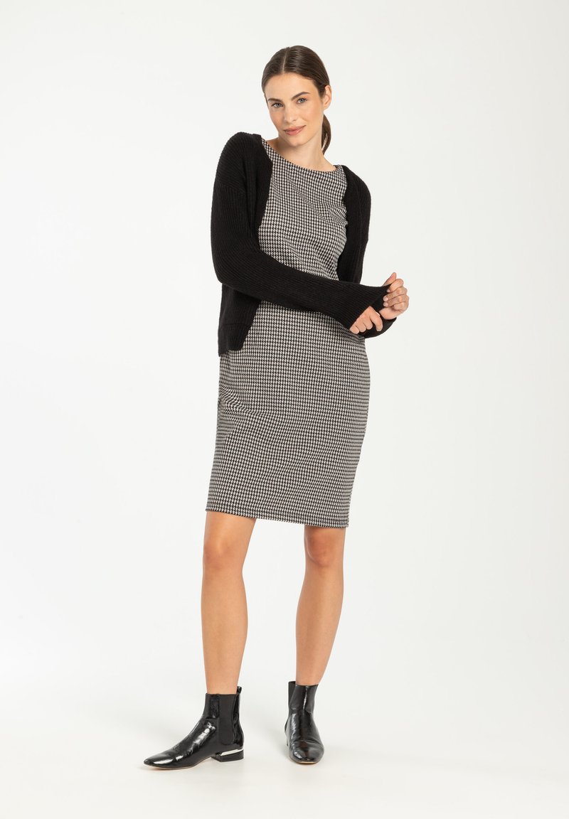 Zwarte cardigan over een zwart-wit gestreepte jurk. De jurk heeft een aansluitende vorm; het model draagt zwarte enkellaarsjes met een glanzende textuur.