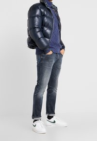Giubbotto imbottito blu navy con finitura lucida, maglione a maglia strutturata, jeans blu sbiaditi e sneakers bianche con dettagli verdi.