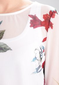 Průsvitný bílý top s barevným květinovým potiskem, včetně červených a modrých květin. Má kulatý výstřih a jemnou texturu.