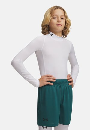 Młoda osoba o długich blond włosach nosi białą dopasowaną bluzkę z długim rękawem i zielone sportowe spodenki, stoi pewnie z rękami na biodrach.