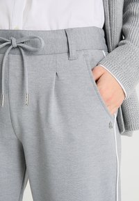 Hellgraue Jogginghose aus weichem Stoff, mit einem Kordelzugbund, seitlichen Taschen und weißen Akzenten entlang der Taschen.