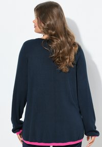 Femme avec de longs cheveux bruns ondulés portant un pull bleu marine avec des bordures roses, vue de dos sur un fond blanc.