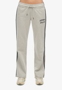 Lichtgrijze sweatpants met een trekkoord in de taille, voorzien van zwarte zijstrepen en het logo "Superdry ATH.JP." op de voorkant.