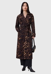 Trench coat con stampa leopardata marrone e nera, con vita cintrata, ampie alette e tasche laterali; abbinato a stivali neri con tacco e a una borsa.