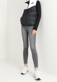 Kvinna i svart och vit Nike puffärmar, svart långärmad tröja, grå skinny jeans och vita sneakers som står mot en vit vägg.