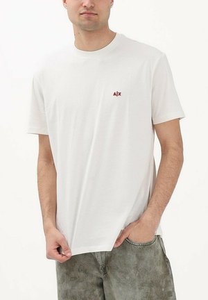 Giovane uomo con maglietta bianca a maniche corte senza disegni con piccolo logo rosso A|X sul petto, jeans grigi slavati, in piedi con una mano in tasca.