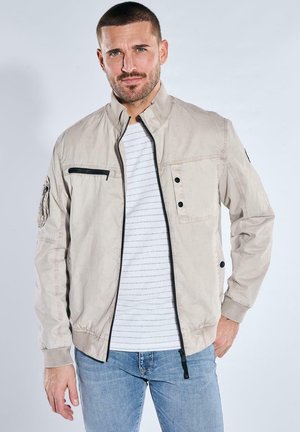 Man wearing beige zip-up jacket over white striped shirt and blue jeans, standing against plain light background.