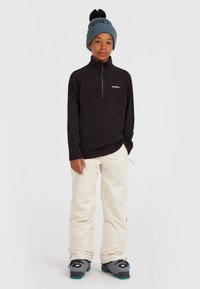 Zwarte fleece hoodie met ritssluiting en een logo, gecombineerd met crèmekleurige sneeuwbroeken. Aangevuld met een lichtblauwe gebreide beanie met een donkere pompon.