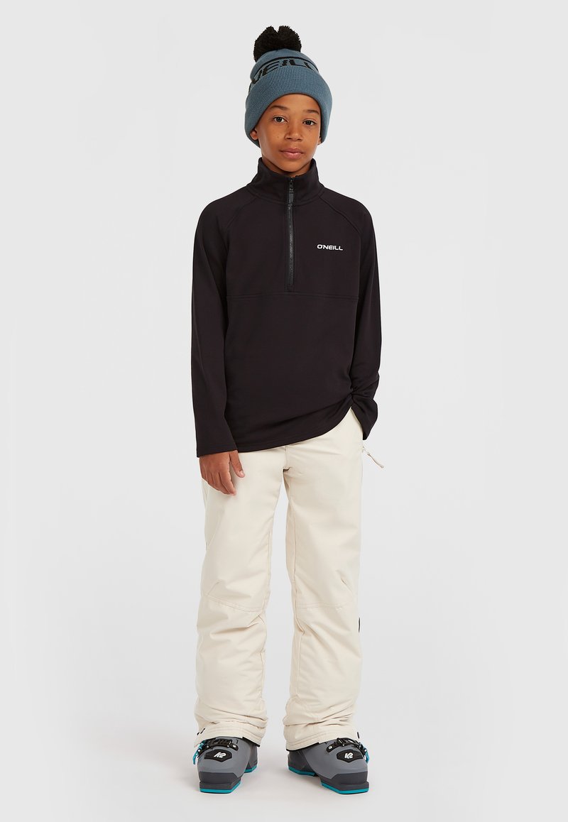 Zwarte fleece hoodie met ritssluiting en een logo, gecombineerd met crèmekleurige sneeuwbroeken. Aangevuld met een lichtblauwe gebreide beanie met een donkere pompon.