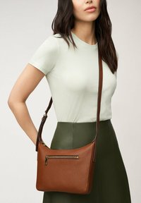 Sac en cuir marron avec une texture lisse, une poche avant zippée et une longue bandoulière réglable. Le design est minimaliste et pratique.