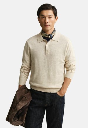 Man die een beige poloshirt met lange mouwen draagt, een donkere spijkerbroek, een patroon sjaal om de nek, een bruine jas in de linkerhand houdt, rechterhand in de zak, neutrale achtergrond.