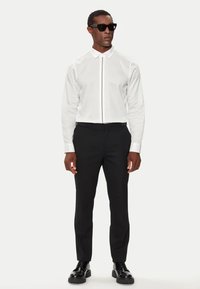 Camicia bianca a maniche lunghe con placket nascosto, abbinata a pantaloni neri su misura e occhiali da sole neri. Scarpe nere lucide completano il look.