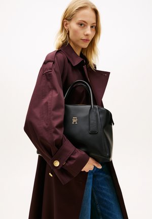 Črna usnjena torbica s strukturiranim dizajnom, kratkimi ročaji, zlatim logotipom in gladko teksturo, držana ob bordo plašču.