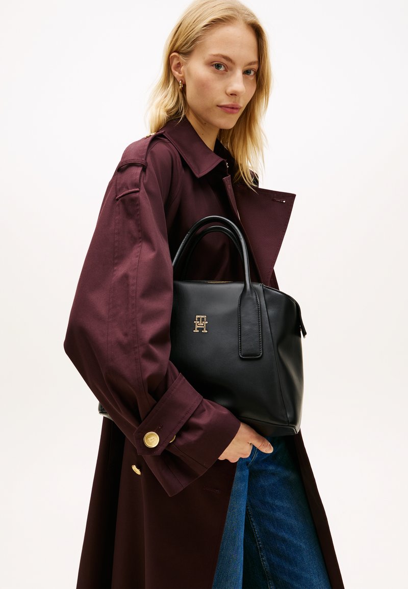 Μαύρη δερμάτινη τσάντα με δομημένο σχέδιο, με κοντά χερούλια, χρυσό λογότυπο και λεία υφή, κρατημένη πάνω σε μπορντό παλτό.
