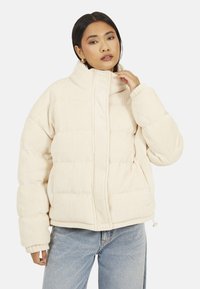 Kremowa pikowana kurtka o prążkowanej fakturze, z wysokim kołnierzem i przednim zamkiem błyskawicznym. Posiada duże kieszenie i elastyczne mankiety. Widoczne dżinsy.