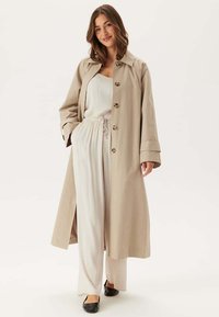 Beige trenchcoat med krage, knäppning framtill, långa ärmar och sidsprickor, bärs över en ljus jumpsuit och platta skor.