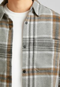 Camisa a cuadros gris hecha de una mezcla de lana, con broches negros, un bolsillo en el pecho y acentos en naranja y blanco en el patrón. Debajo lleva un suéter blanco.