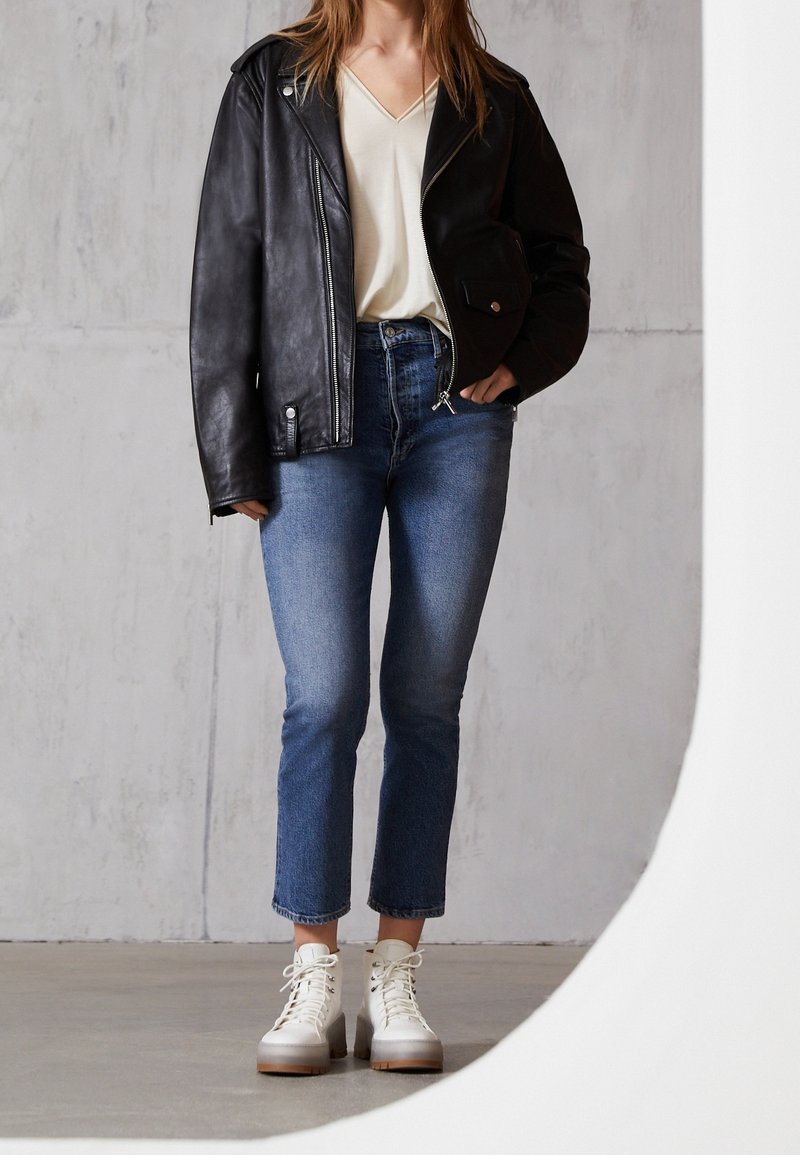 Personne portant une veste en cuir noire, un t-shirt blanc col en V, un jean court bleu et des bottines blanches à lacets, debout à l'intérieur.