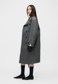 Manteau en laine gris avec grand col, boutons croisés et poches latérales. Gants et chaussettes bleu marine complètent le look. Chaussures plates et pointues visibles.