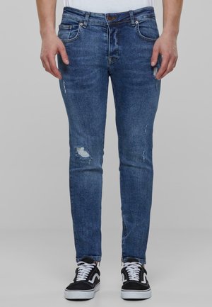 Jean skinny en denim bleu moyen avec des détails déchirés, présentant une taille mi-haute et un design à cinq poches, associés à des baskets noires.