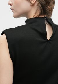 Sieviete ar īsiem brūniem matiem, tērpusies melnā bezplecu topā ar atvērumu aizmugurē, kas atklāj nelielu kakla tetovējumu.