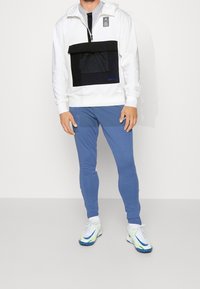 Sudadera blanca con capucha y un bolsillo de malla negro, combinada con pantalones ajustados azul claro y zapatillas blancas con acentos verdes y azules.