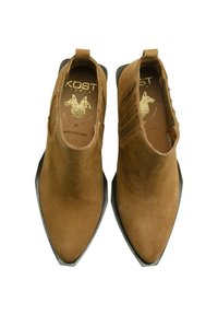 Bottines en daim marron avec des orteils pointus, des accents élastiques sur les côtés et une semelle en caoutchouc noire. Logo doré visible sur la semelle intérieure.