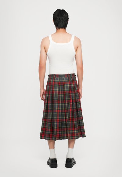 Ein Tartankilt in den Farben Rot, Grün und Gelb. Gefaltetes Design, taillierter Bund, getragen mit einem weißen Tanktop und schwarzen Schuhen.