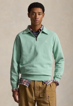 Άνδρας με πουλόβερ σε μέντα πράσινο με φερμουάρ στο ένα τέταρτο πάνω από καρό πουκάμισο και μπεζ παντελόνι cargo, στέκεται με το ένα χέρι στην τσέπη, μπροστά σε απλό φόντο.