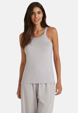 Vrouw met lang bruin haar, gekleed in een lichtgrijze mouwloze tanktop en bijpassende losse broek, staande tegen een effen achtergrond.