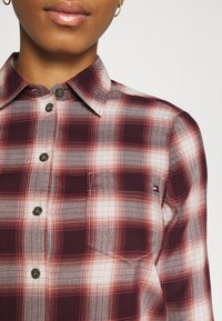 Personne portant une chemise à boutons à carreaux bordeaux et blancs avec une poche poitrine et un petit logo rectangulaire, montrant une partie des lèvres et du cou.