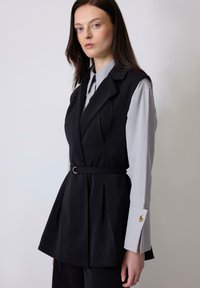 Black tailored vest with a belted waist, pleated design, and a structured collar, layered over a light gray button-up shirt with gold accents.
