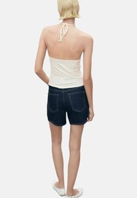 Top bianco a halter con lacci sulla schiena, abbinato a pantaloni shorts in denim blu scuro con tasche posteriori e cuciture a contrasto, indossato con scarpe bianche.