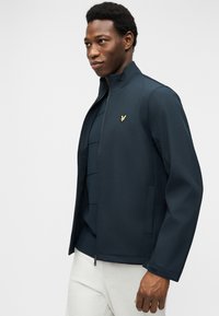 Νέος άνδρας που φοράει ένα σκούρο μπλε μπουφάν με φερμουάρ και χρυσό έμβλημα πουλιού, λευκό παντελόνι και σκούρο πουκάμισο με στρώσεις από μέσα, στέκεται μπροστά σε απλό φόντο.