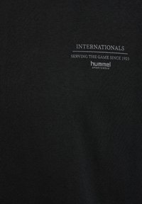 Čierne bavlnené tričko s bielym potlačou textu: "INTERNATIONALS," "SERVING THE GAME SINCE 1923," a "hummel sportswear." Hladký materiál.