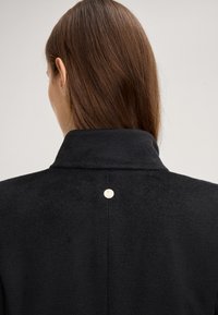 Zwarte jas met een zachte textuur, een hoge kraag en een opvallend zilverkleurig accent op de achterkant nabij de halslijn.