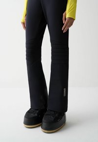 Persona che indossa pantaloni da sci neri imbottiti con il logo "COLMAR" e stivali da neve neri, in piedi su uno sfondo bianco uniforme.