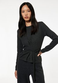 Top de manga larga gris oscuro con diseño de peplum, detalle de cintura atada y escote redondo. La tela parece suave y con textura.