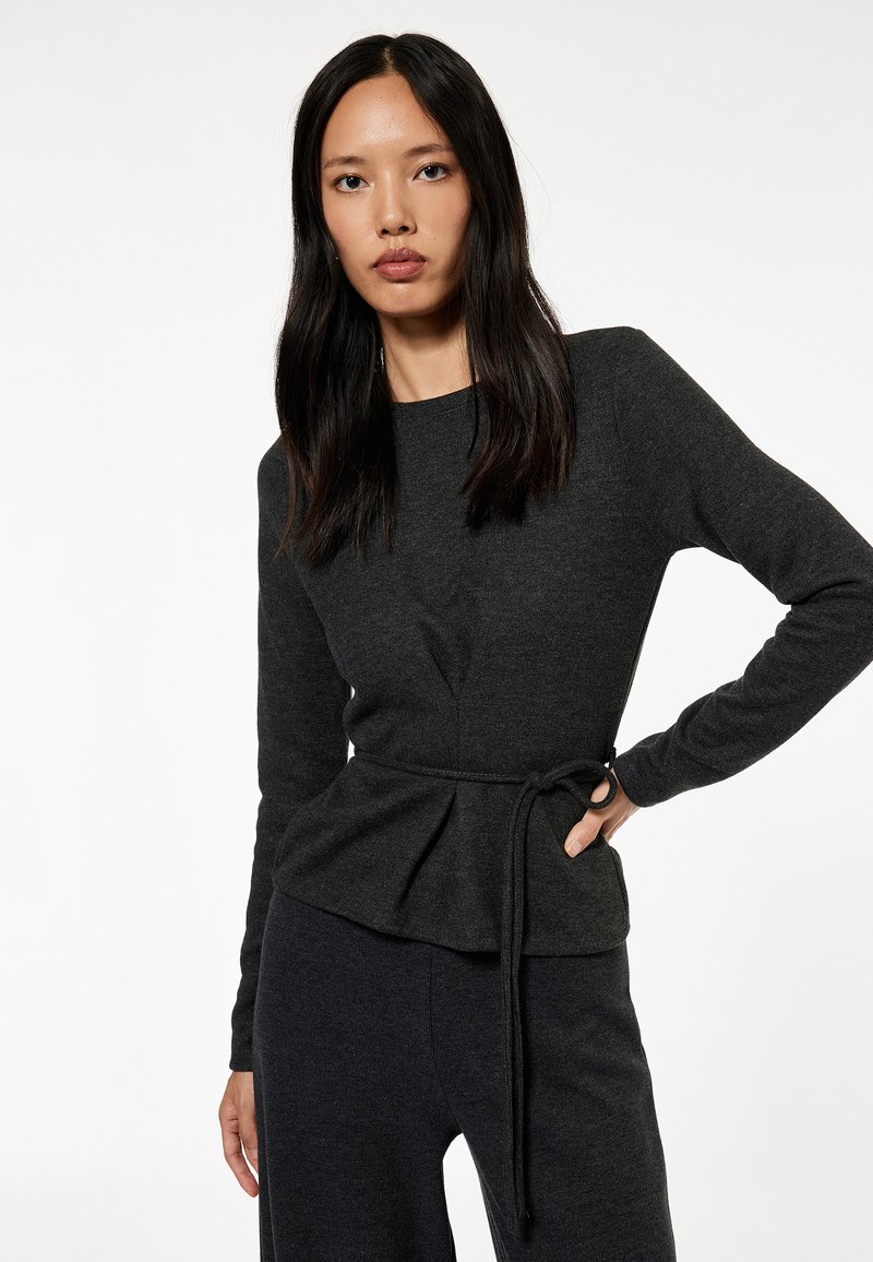 Top de manga larga gris oscuro con diseño de peplum, detalle de cintura atada y escote redondo. La tela parece suave y con textura.