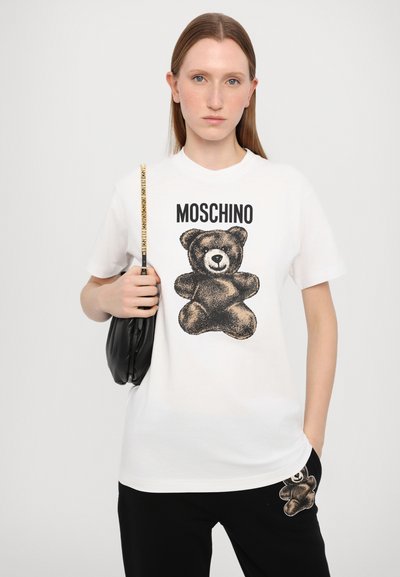 Hvit bomull t-skjorte med grafikk av en svart bjørn og teksten "MOSCHINO". Modellen holder en svart pose og har på seg sorte bukser med bjørnedetaljer.