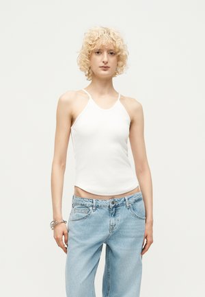 Νεαρό άτομο με κατσαρά ξανθά μαλλιά που φοράει λευκό αμάνικο τοπ και ανοιχτό μπλε τζιν μπροστά σε απλό ανοιχτό φόντο.