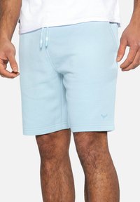 Shorts décontractés bleu clair avec taille à cordon et poches portés par une personne se tenant devant un fond uni.