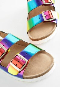 Regnbågsfärgade sandaler har två metalliska remmar med silverfästen, en strukturerad tan-fotbädd och en vit gummisula.