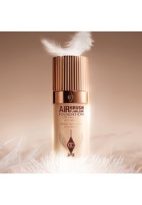 Airbrush Flawless Foundation in a sleek glass bottle with a ribbed golden cap. Features text in brown and gold. Soft feather accent nearby.