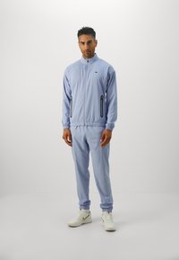 Mand står iført en lyseblå zip-up træningsjakke og matchende bukser med hvide Nike sneakers foran en ensfarvet grå baggrund.