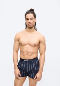 Homme torse nu avec des dreadlocks portant un boxer rayé bleu marine avec une ceinture "BOSS", debout devant un fond clair uni.