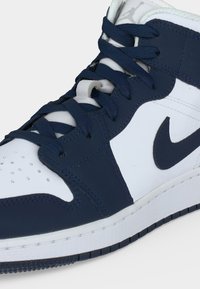 Αθλητικό παπούτσι Nike Air Jordan με υψηλή γλώσσα, λευκό δέρμα, ναυτικό μπλε επικάλυψη, ταίριασμα κορδονιών και εμφανές λογότυπο Jumpman στη γλώσσα.